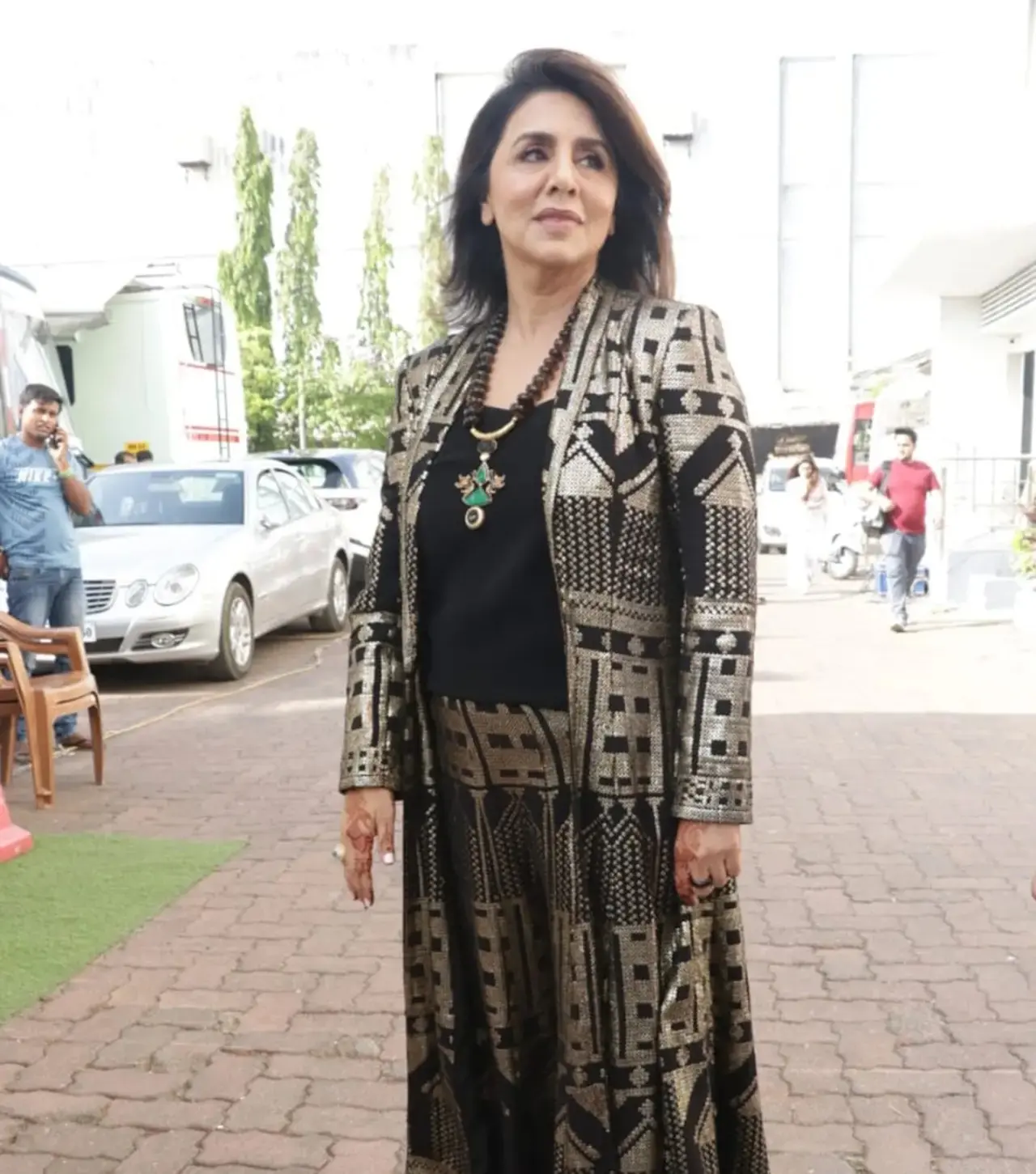 Neetu Kapoor looks absolutely striking in this modern ethnic ensemble, featuring a dramatic floor-length open jacket over a sleek black base. The jacket is adorned with bold, geometric gold and silver sequin patterns.