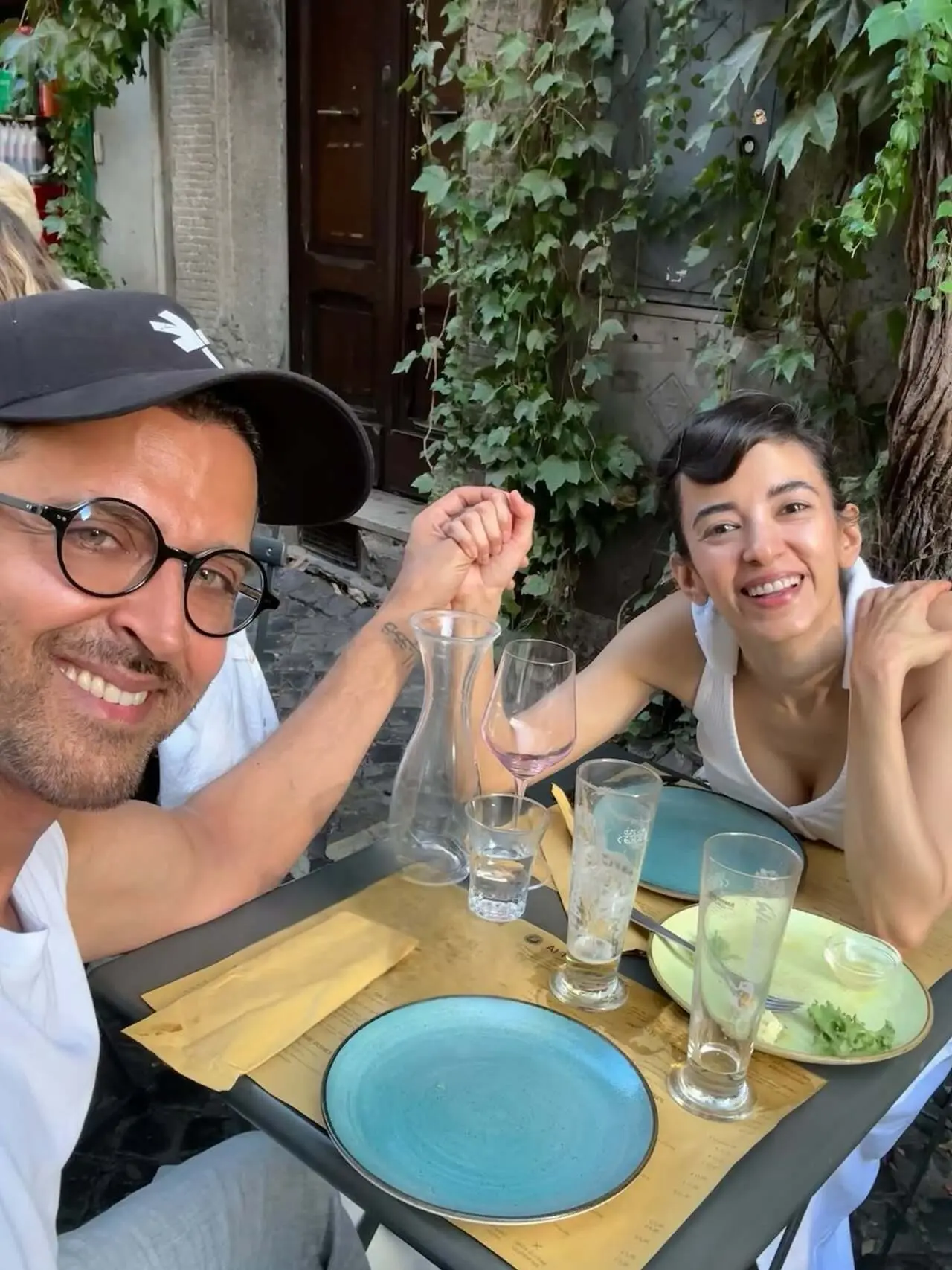Hrithik Roshan and Saba Azad beam at the camera while holding hands across an outdoor restaurant table during a relaxed outing
