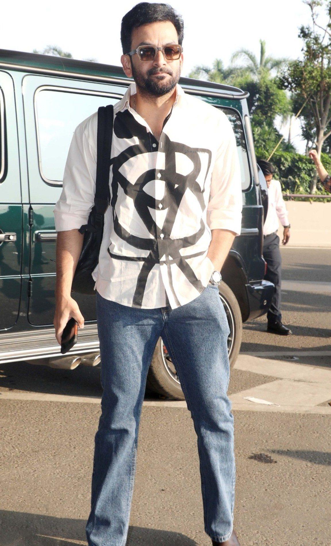 Prithviraj Sukumaran was recently spotted at the airport keeping it sharp and stylish in a graphic white shirt paired with classic blue jeans and brown shoes. Prithviraj Sukumaran was recently spotted at the airport keeping it sharp and stylish in a graphic white shirt paired with classic blue jeans and brown shoes.