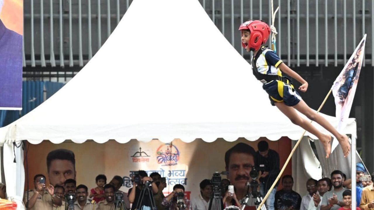 A young participant soars mid-air during Dahi Handi celebrations — helmet on, flag in tow, crowd roaring below A young participant soars mid-air during Dahi Handi celebrations — helmet on, flag in tow, crowd roaring below