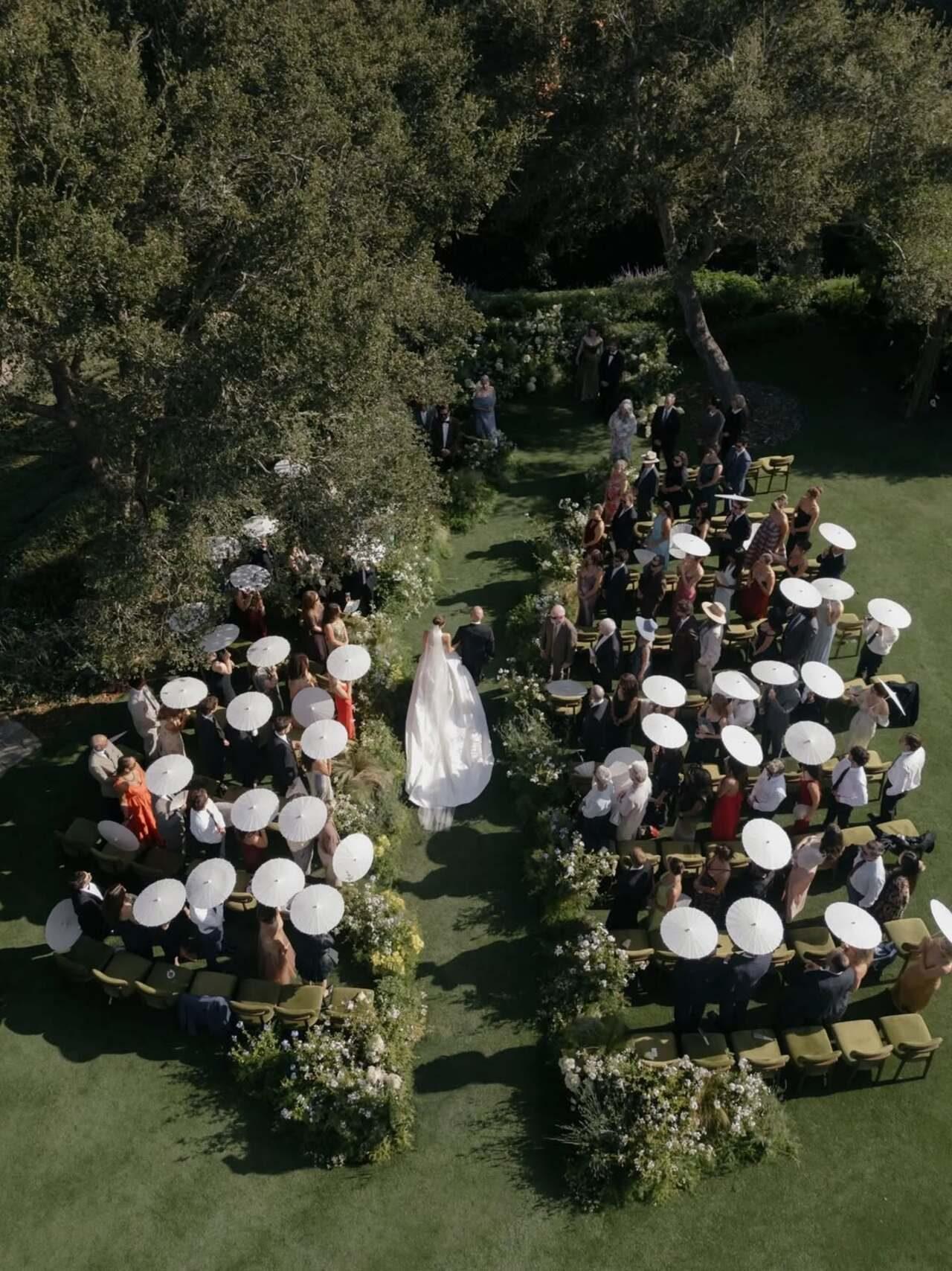 An overhead shot of the outdoor ceremony set on a lush green lawn lined with floral arrangements. Guests seated on both sides hold white parasols to shade themselves as the bride processes down the aisle.
