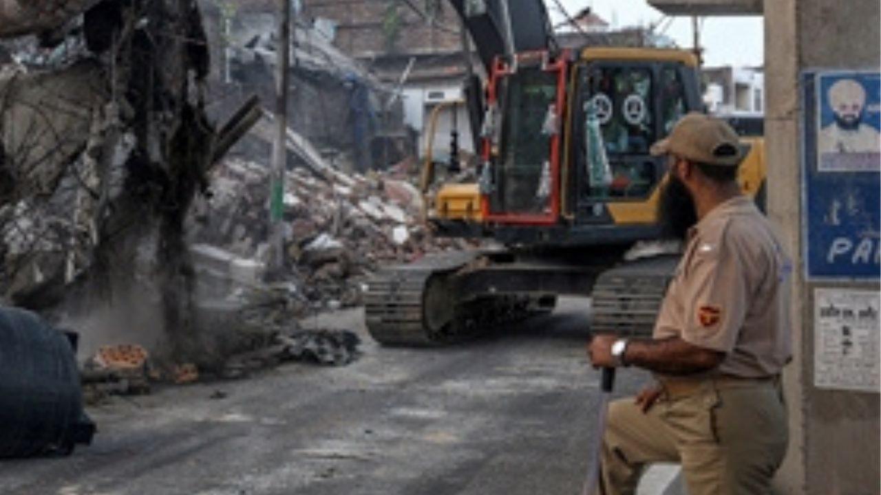 A police official stands guard as a demolition drive being carried out by district administration at Nai Basti area, in Jammu A police official stands guard as a demolition drive being carried out by district administration at Nai Basti area, in Jammu