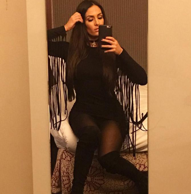 Nikki Bella looks stylish in a black dress as she clicks a selfie while seated on a bed.